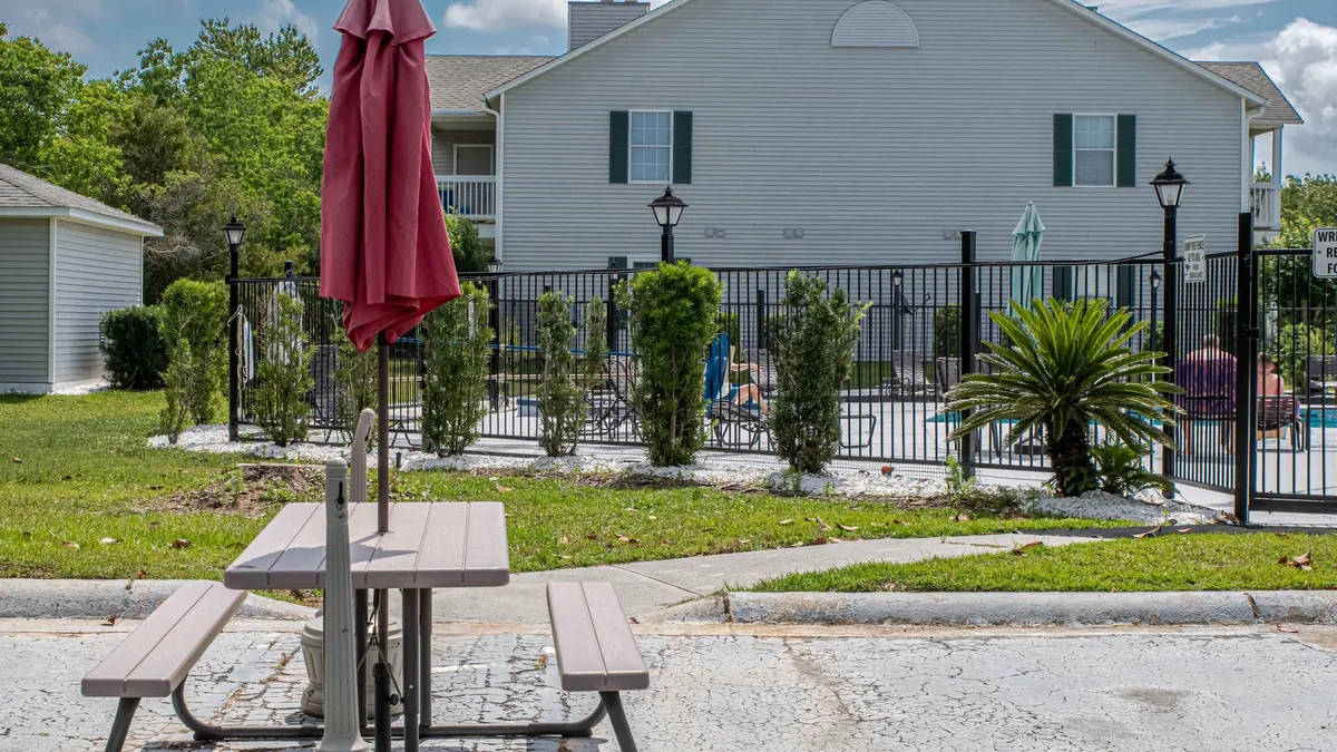 A sunlit swimming pool area with a picnic table shaded by a large umbrella, surrounded by landscaped greenery and a secure fenced border, inviting small groups for poolside meals or relaxation.