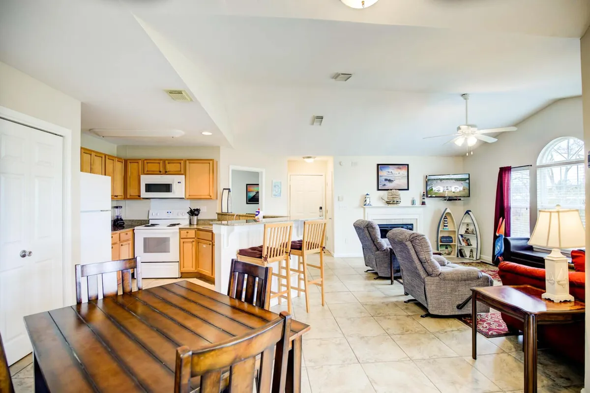 A spacious, open-concept room featuring a dining table, kitchen cabinetry, and living room seating. The layout visually connects cooking, dining, and lounging areas, complemented by stylish decor and a decorative shelf.