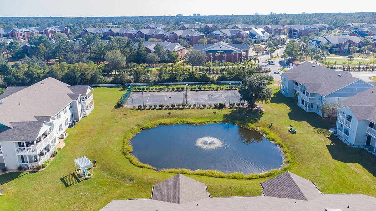 A serene pond with a central fountain is surrounded by lush greenery and recreational areas. A walking path circles the pond, and nearby tennis courts hint at opportunities for outdoor activity. The setting is peaceful, inviting for both social gatherings and solitary strolls.