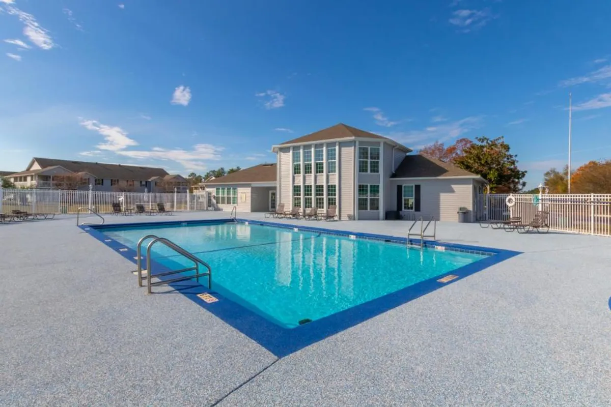 An inviting swimming pool area with lounge chairs and a clubhouse nearby. The pool is bordered by palm trees and landscaping, and the calm water glistens under the sun, inviting guests to swim, sunbathe, or socialize.