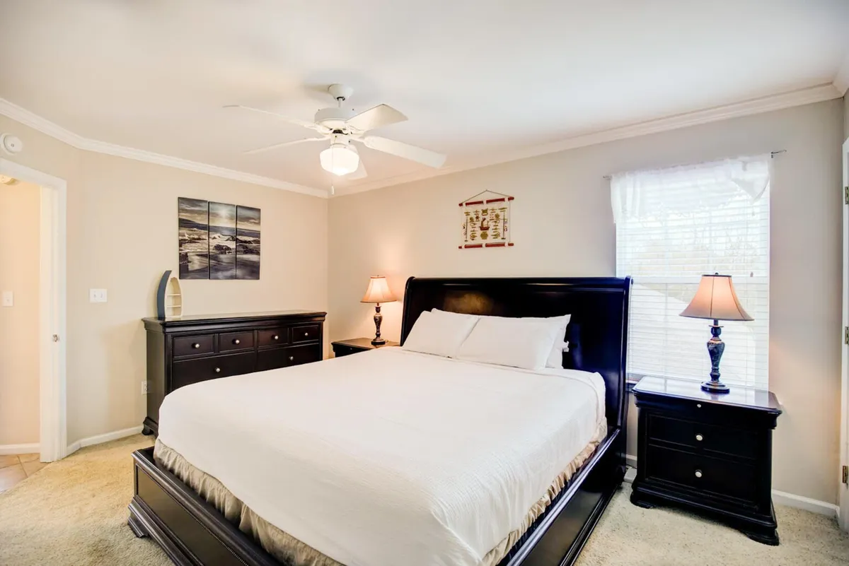 A tranquil master bedroom featuring a king-size bed with plush linens, matching nightstands with lamps on either side, and decorative artwork above the headboard. Soft natural light filters through the window, creating an inviting and restful ambiance.