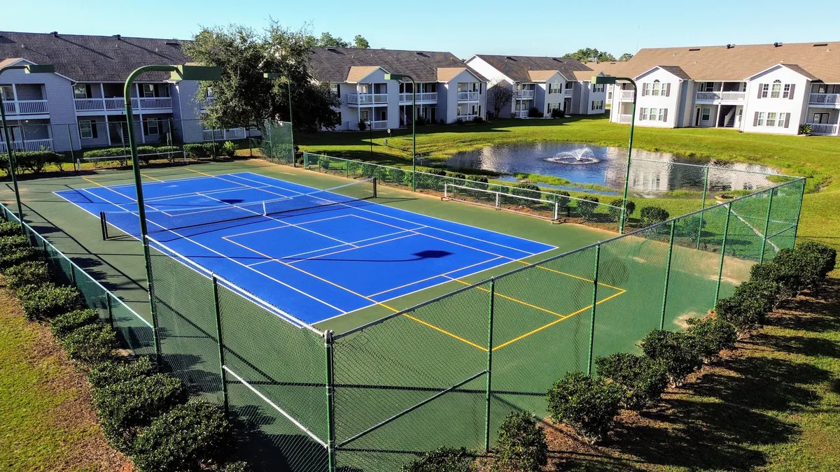 A well-maintained exterior with tennis courts adjacent to a tranquil pond and surrounded by lush greenery and apartment buildings. The courts are ready for friendly matches and foster a sense of community activity.
