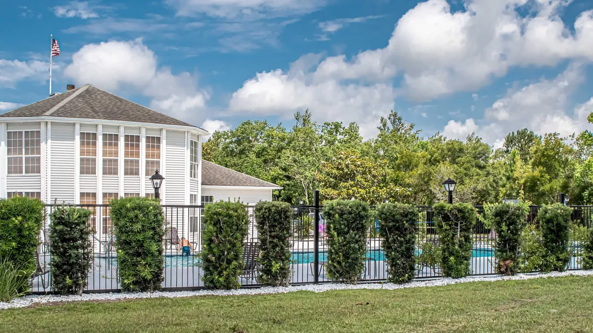 This image shows a sparkling swimming pool surrounded by a fenced area, lush landscaping, and an adjacent building structure. The inviting blue water, ample poolside space, and vibrant greenery create a resort-like atmosphere perfect for socializing and relaxation.
