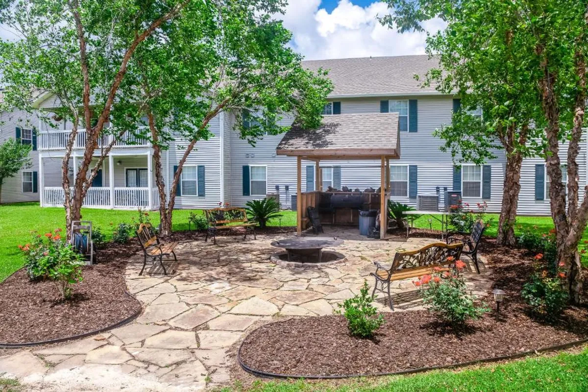 A welcoming stone patio area featuring a grill, fire pit, comfortable outdoor seating, and vibrant flower beds. The setting suggests a space perfect for communal barbecues, social gatherings, and relaxed evenings outdoors.
