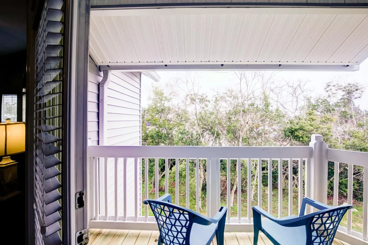 A peaceful private balcony with blue outdoor chairs, white railing, and a view of lush trees and greenery beyond. The setting evokes a sense of tranquility and an ideal spot for morning coffee or watching the sunset.