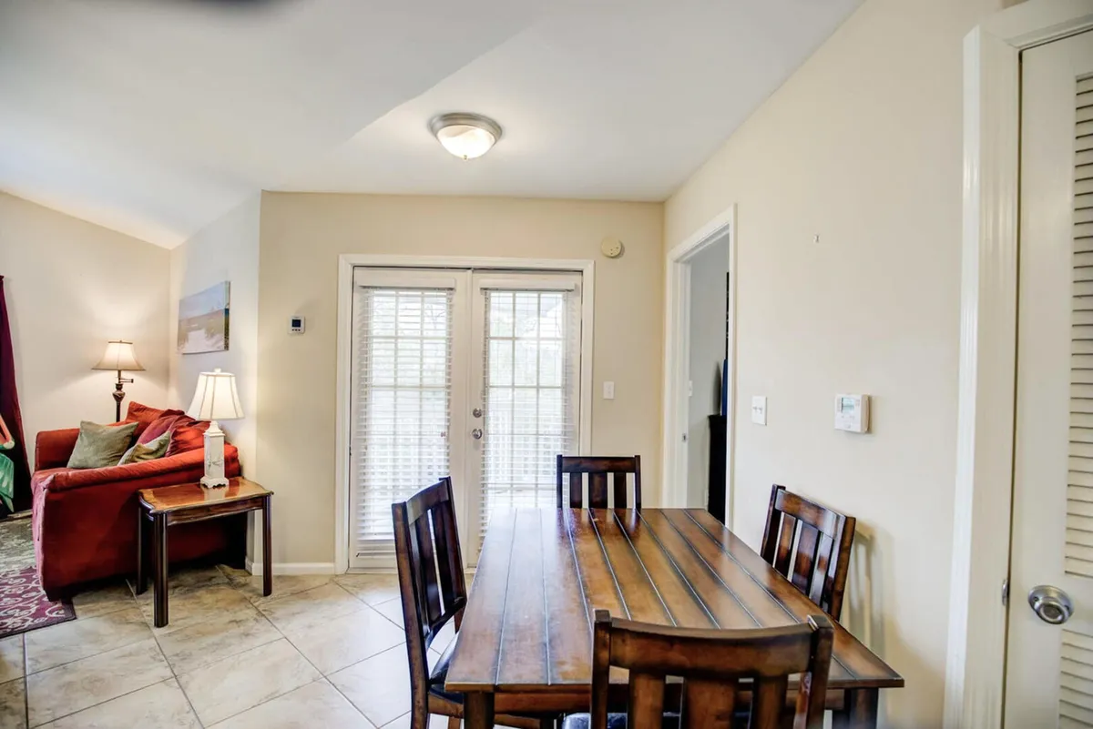 This image presents a warm, inviting living and dining area featuring a wooden dining table with four chairs, a cozy red sofa with cushions, a side table with a lamp, and French doors with blinds that allow natural light to pour in. The French doors likely lead to the private balcony, visually connecting the indoor and outdoor spaces.
