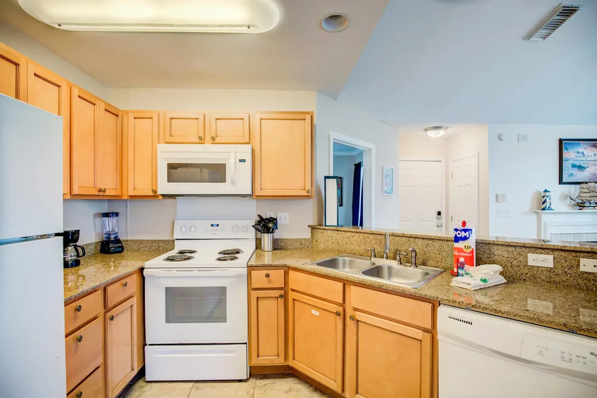 A functional, inviting kitchen is shown with wooden cabinets, a white electric stove and microwave, double stainless steel sink, and granite countertops. The layout supports easy meal prep and casual dining at the counter.