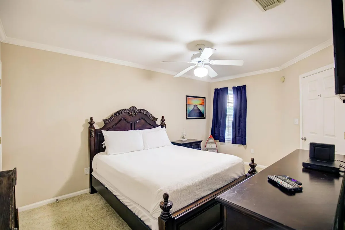 A serene king bedroom featuring an ornate dark wood king-size bed with crisp white linens, a dark wood dresser topped with a TV and remote, a small nightstand with a lamp, and a window dressed with dark blue curtains. The room exudes a tranquil, restful ambiance ideal for winding down after a busy day.