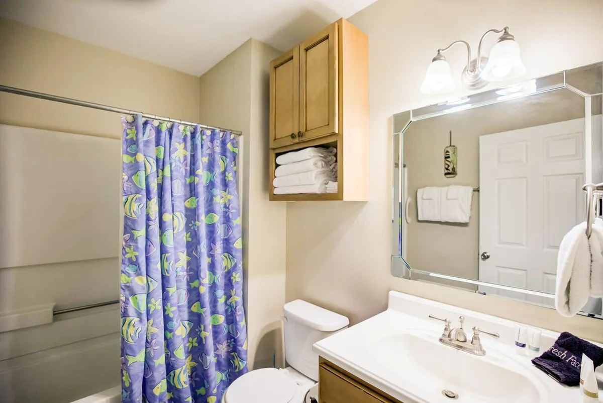 A cheerful bathroom with a colorful fish-themed shower curtain, a white sink and countertop, a wooden cabinet stacked with fresh towels, and a large mirror with a well-lit fixture. The space is both practical and inviting, with a playful touch.