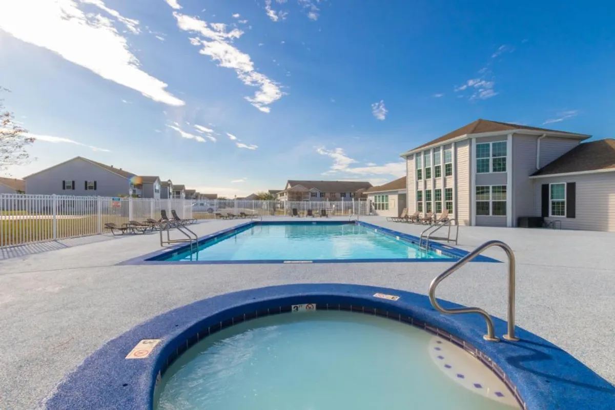 A large outdoor swimming pool and circular hot tub/jacuzzi are featured in the foreground, with a two-story clubhouse and poolside lounge chairs along the fenced perimeter. The environment is relaxing, inviting, and promotes both leisure and social interaction.