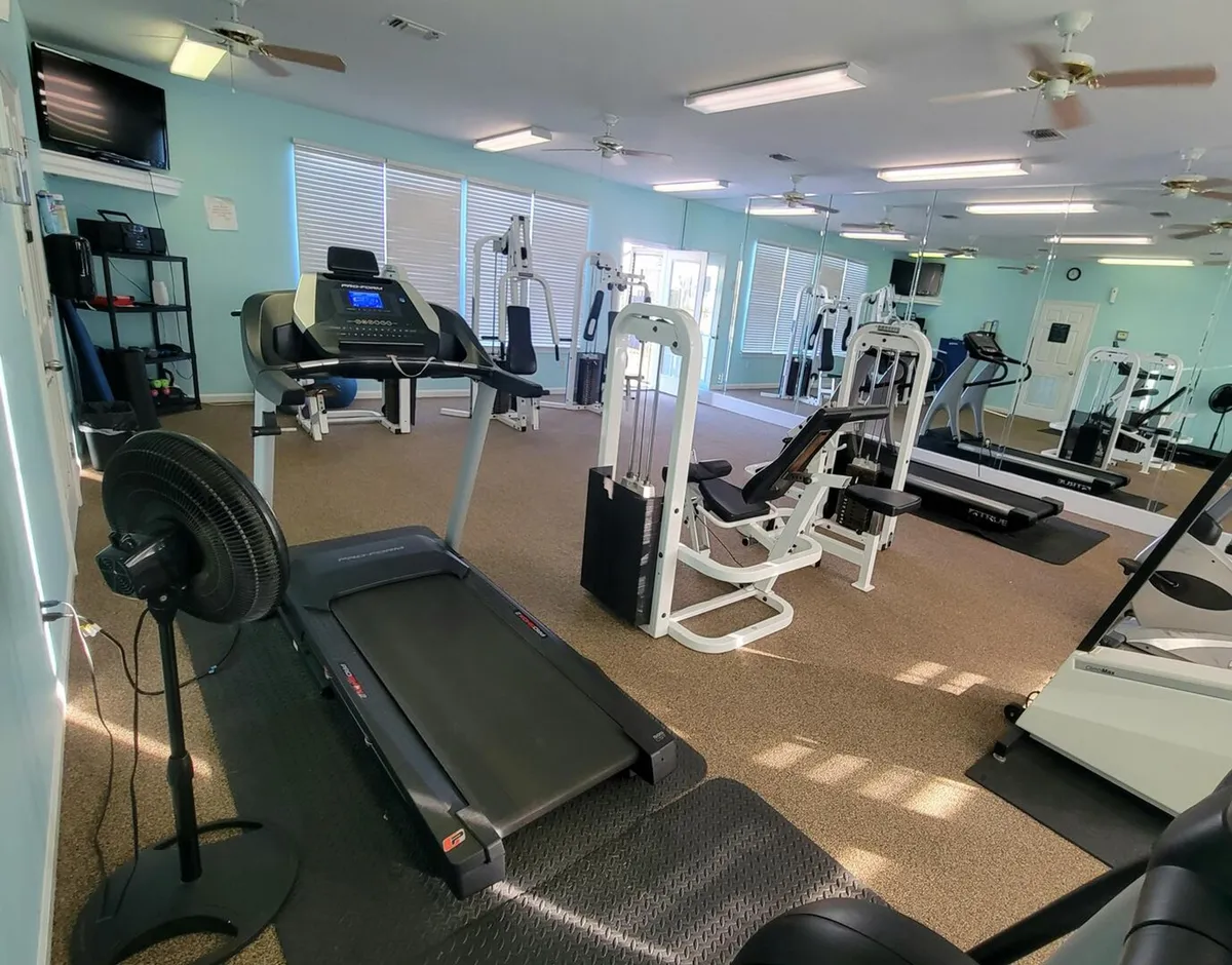 A fitness center interior equipped with a treadmill, weight machines, a large wall mirror, and ceiling fans. The room is bright and designed for both cardio and strength training.