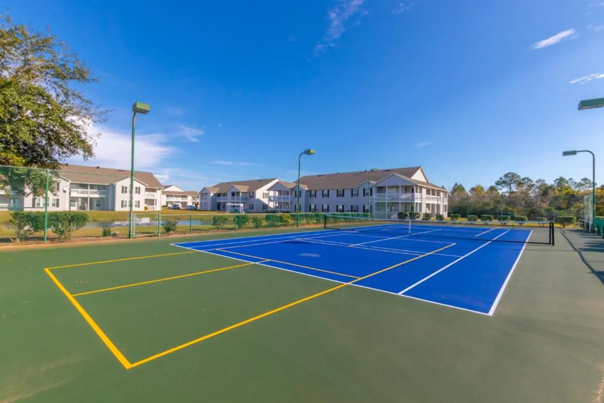 This exterior image showcases a well-maintained blue tennis court bordered by lush landscaping and residential buildings, with tall outdoor lights indicating night play potential. The setting is tranquil yet active, inviting guests to engage in healthy, recreational activities.