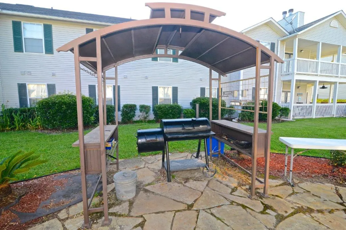 A covered barbecue grill station with a smoker, set on a stone patio beside well-maintained lawns and landscaped grounds. Nearby, residential buildings provide a sense of community and accessibility.