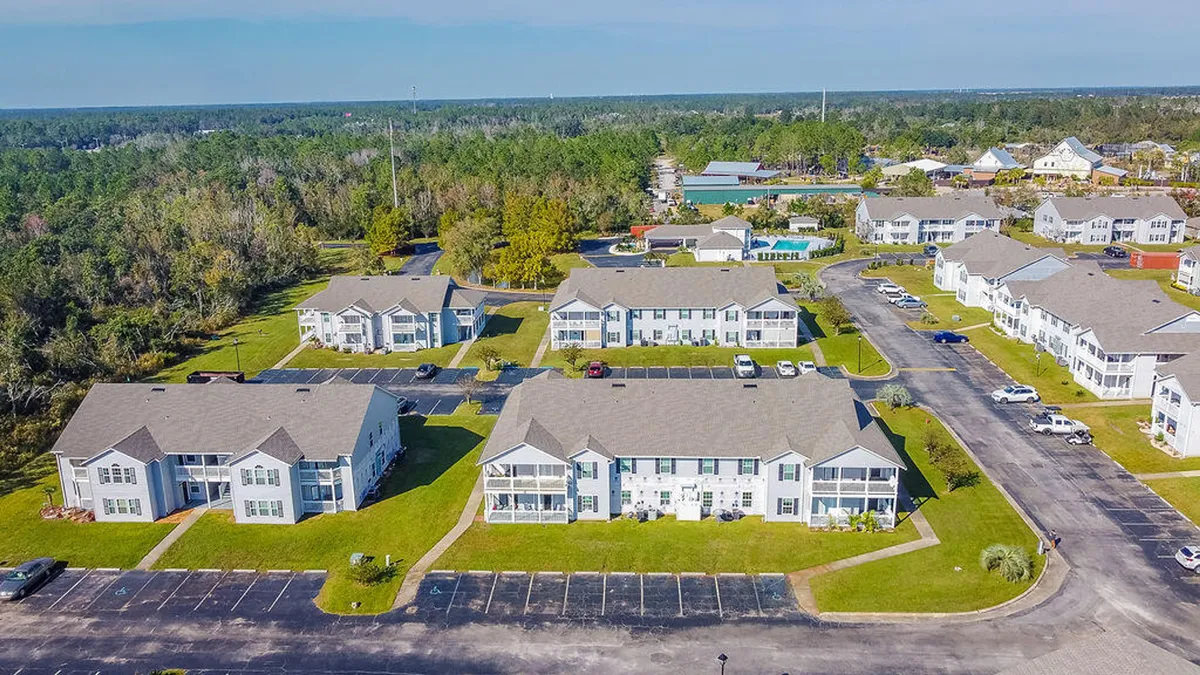 The image features multiple white two-story condo buildings with gray roofs, surrounded by well-kept green lawns and landscaping. Parking areas and a central community pool are visible, set within a peaceful, family-friendly environment.