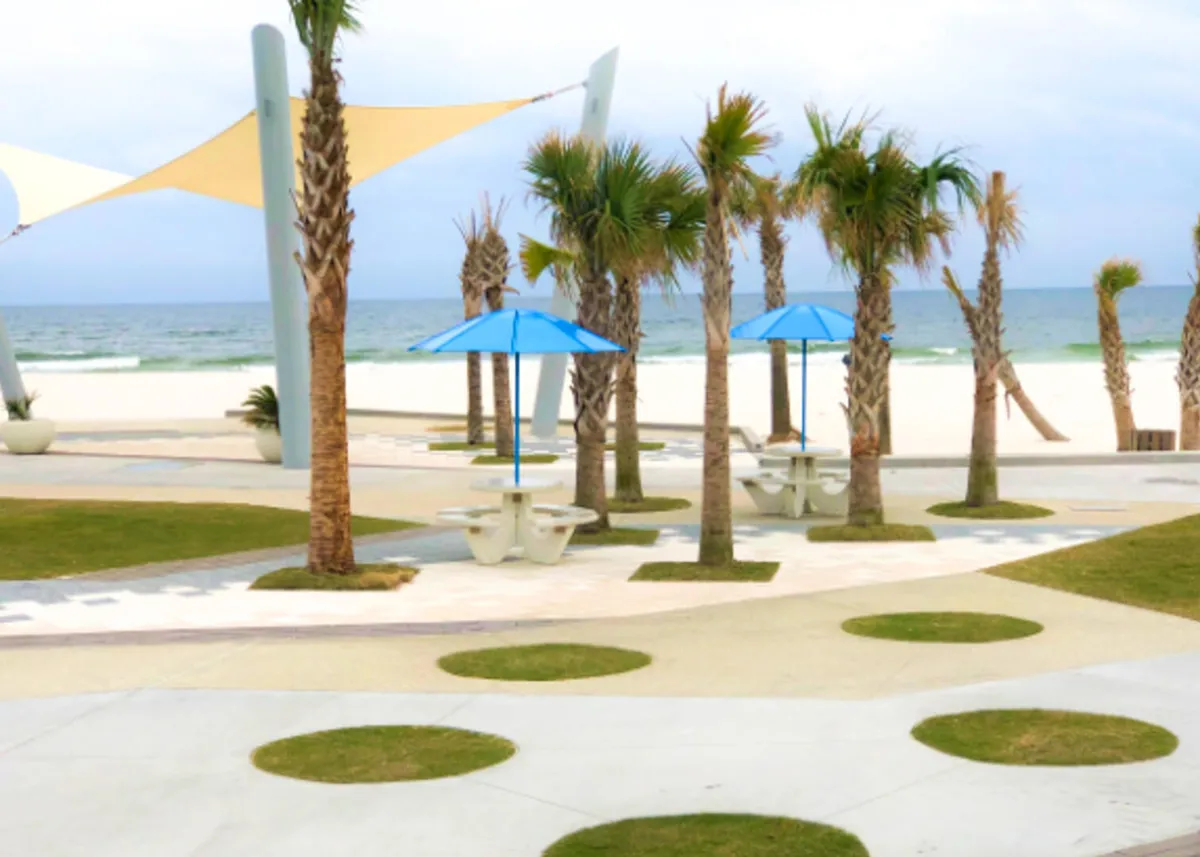 This image captures a beachside area lined with palm trees, blue umbrellas shading picnic tables, and a white sandy beach stretching to calm ocean waves in the background. The mood is relaxed and refreshing, ideal for social gatherings and enjoying the Gulf Coast scenery with friends.