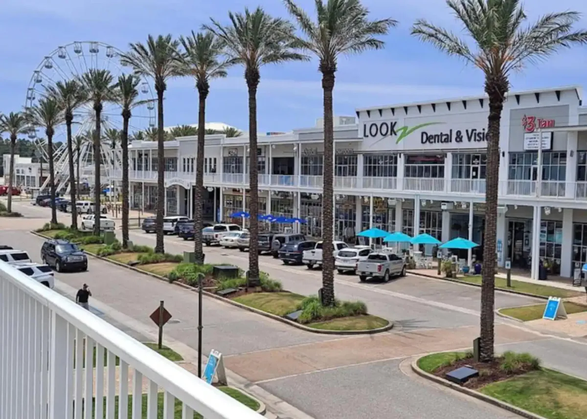 A lively exterior scene with a Ferris wheel in the background, palm trees lining the street, a two-story commercial building with visible signage, and outdoor seating with blue umbrellas. The area evokes a sense of vibrant shopping and dining nearby.